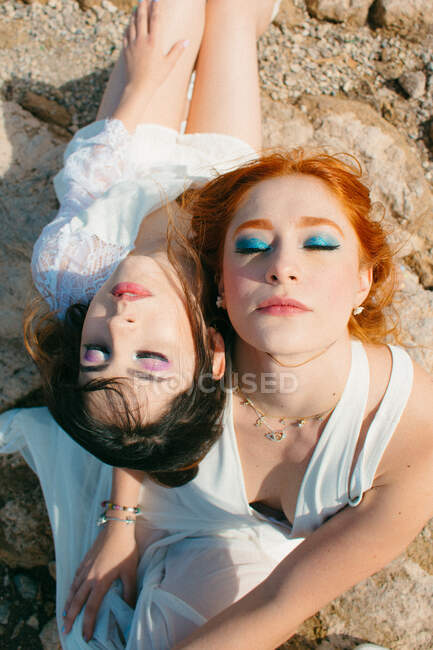 Вид зверху на молоду гомосексуальну пару з закритими очима в білих сукнях, що сидять на нерівній землі на сонячному світлі — стокове фото