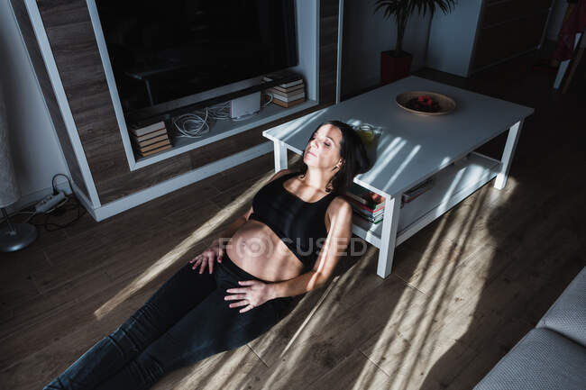 Alto ângulo de tranquila fêmea grávida apoiada na mesa e sentada no chão em casa enquanto desfruta da luz solar com olhos fechados — Fotografia de Stock