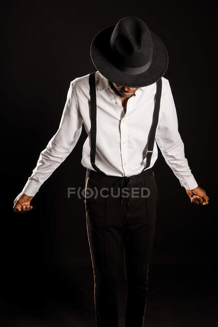 Jovem modelo masculino étnico em chapéu e calças de pé dançando em fundo preto — Fotografia de Stock
