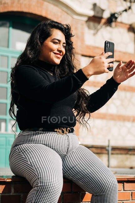 Весела плюс розмір жінка в модному одязі, сидячи на паркані і знімаючи себе на смартфоні в сонячний день у місті — стокове фото