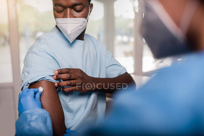 Especialista médico irreconocible en uniforme protector y guantes de látex que desinfecta el brazo con algodón con alcohol para vacunar al paciente afroamericano masculino en la clínica durante el brote de coronavirus - foto de stock