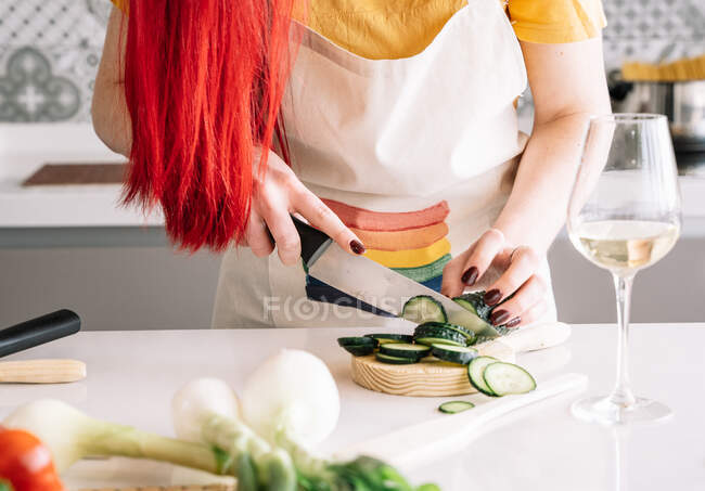 Cultivar homossexual fêmea irreconhecível cortando pepino fresco com faca enquanto prepara alimentos saudáveis na mesa da cozinha com copo de vinho — Fotografia de Stock