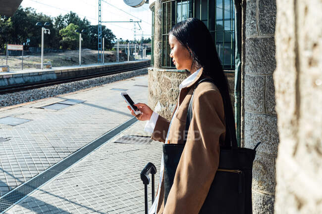 Side view of focused ethnic female traveler standing with suitcase on platform at railroad browsing on smartphone — Stock Photo