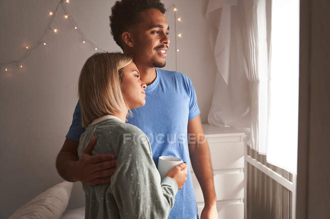 Seitenansicht eines lächelnden multiethnischen Paares im Pyjama, das sich morgens umarmt, während es im Schlafzimmer steht und aus dem Fenster schaut — Stockfoto