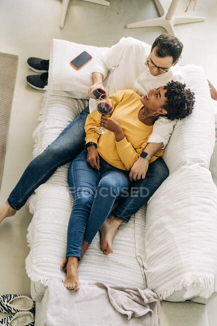 Conteúdo casal multirracial que arrefece no sofá em casa com vinho tinto em óculos enquanto desfruta de fim de semana em casa e olha um para o outro — Fotografia de Stock