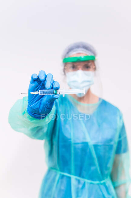 Médecin flou méconnaissable dans un masque protecteur et gants en latex avec flacon de vaccin contre le coronavirus et seringue montrant à la caméra alors qu'il se tenait debout dans la chambre d'hôpital — Photo de stock