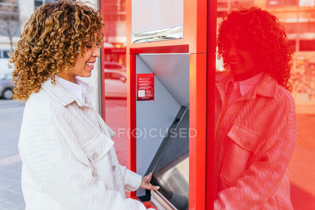 Side view of smiling African American female using ATM terminal and withdrawing cash while standing on city street — Stock Photo