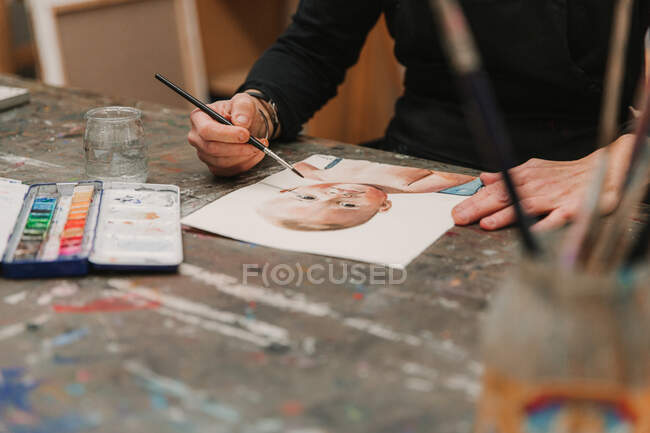 Female artist in apron painting with watercolors on paper while sitting at table in creative workshop — Stock Photo
