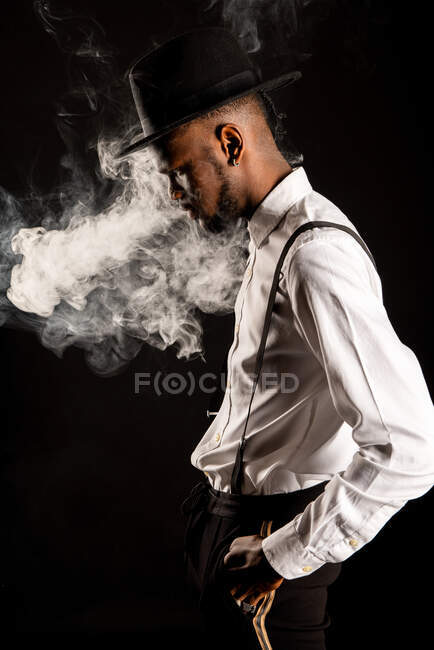 Vista lateral de varón masculino afroamericano en camisa blanca y sombrero exhalando vapor mientras fuma cigarrillo electrónico - foto de stock