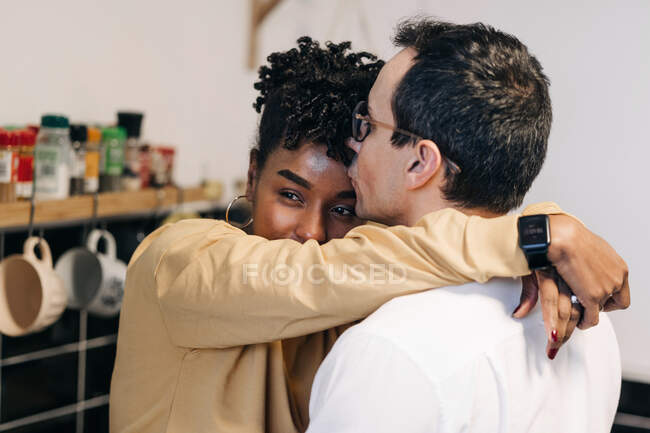 Vista lateral do casal multirracial amoroso abraçando suavemente enquanto está na cozinha em casa — Fotografia de Stock