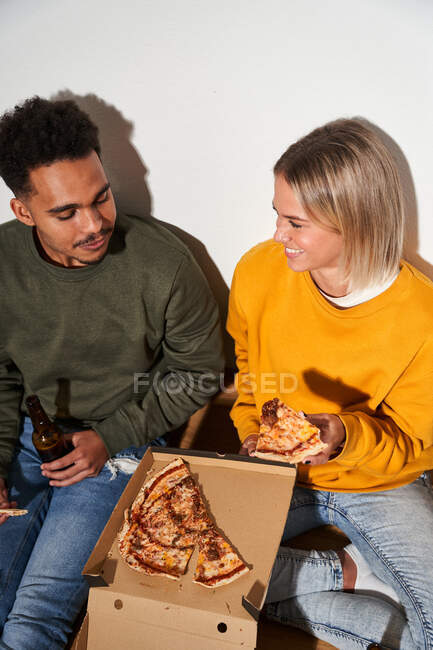 Couple multiethnique joyeux assis sur le sol à la maison tout en mangeant de délicieuses pizzas et en buvant de la bière ensemble — Photo de stock