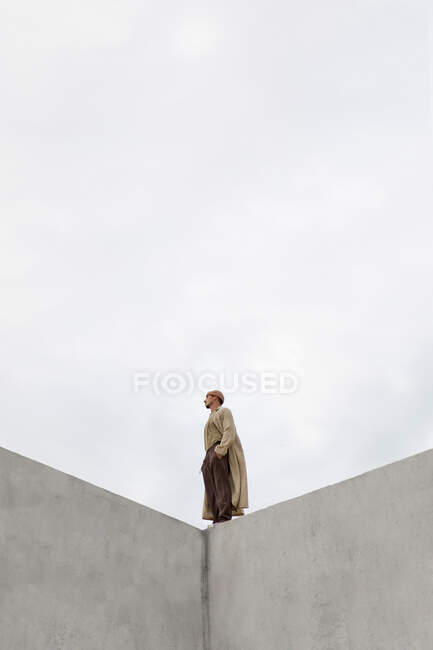 Вид збоку самотнього чоловіка, що стоїть з руками в кишенях на бетонній будівлі проти сірого неба і дивиться вниз — стокове фото