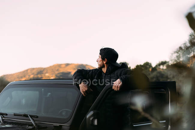 Side view of an adventurer leaning on the door of an off-road car while looking away before starting the journey — Stock Photo