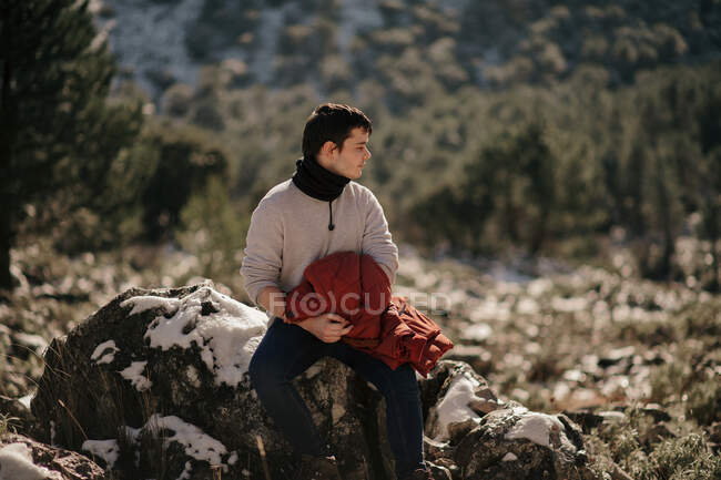 Junge männliche Touristen in warmer Kleidung betrachten die Natur, während sie auf einem Stein gegen den Berg sitzen und im Sonnenlicht wegschauen — Stockfoto