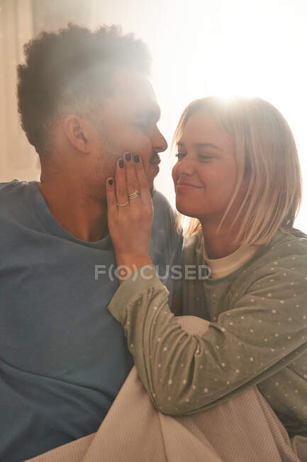Happy woman touching face of black man while sitting on bed after awakening and enjoying sunny morning — Stock Photo