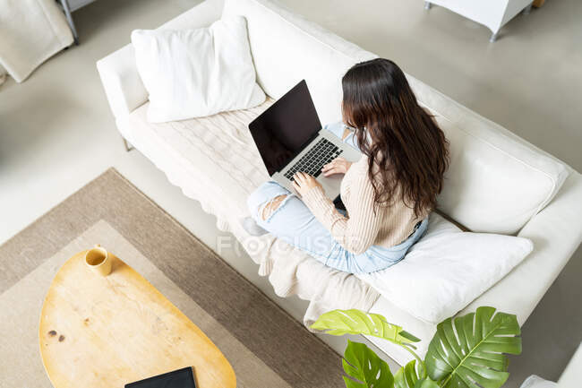 From above back view of anonymous female typing on netbook with black screen while sitting on couch in house — Stock Photo