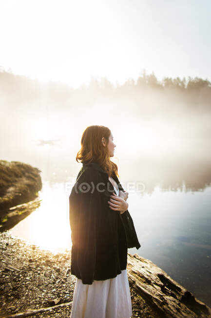 Mujer de pie vestida con un vestido blanco y una chaqueta sobre ella en una roca mirando a un lago en un día de niebla - foto de stock