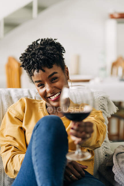 Задоволена афроамериканка сидить на дивані зі склом червоного вина і відпочиває вдома, дивлячись на камеру. — стокове фото