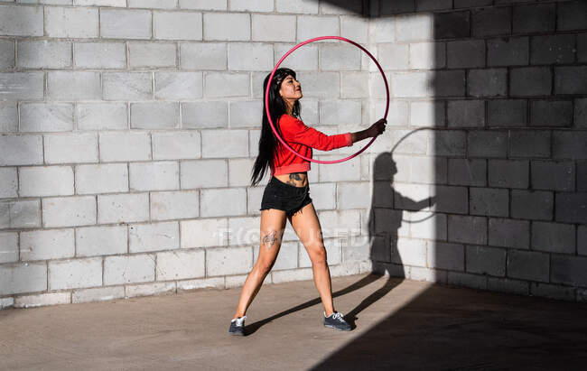 Young tattooed woman in activewear twirling hula hoop while dancing against brick walls with shadows and looking forward in sunlight — Stock Photo