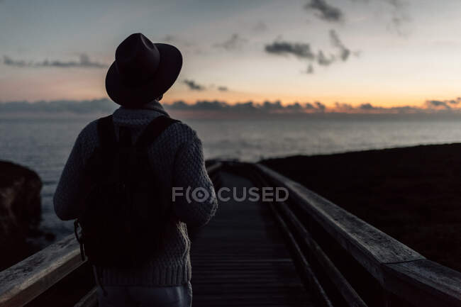 Человек в рюкзаке и шляпе стоит и смотрит на море — стоковое фото