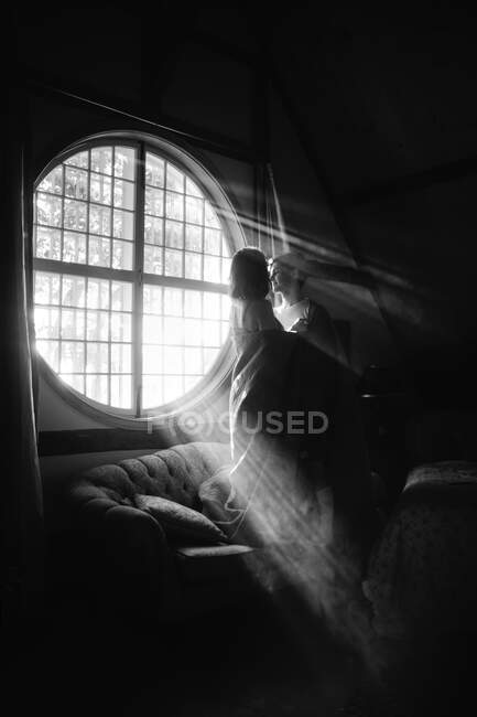 Анонімний чоловік обіймає дівчину текстилем на дивані, дивлячись один на одного на круглому вікні в сонячному світлі — стокове фото