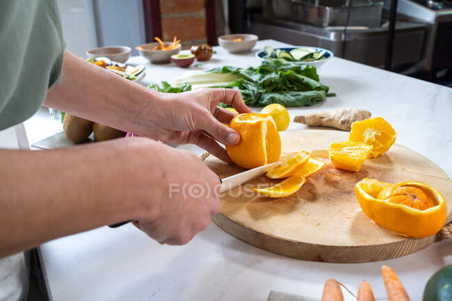 Ernte unkenntlich Weibchen schälen reife saftige Orangen mit Messer auf Holzschneidebrett am Küchentisch — Stockfoto