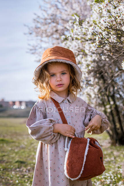 Entzückendes kleines Mädchen in Kleid und Hut steht am Baum mit blühenden Blumen und blickt in die Kamera im Frühlingsgarten — Stockfoto