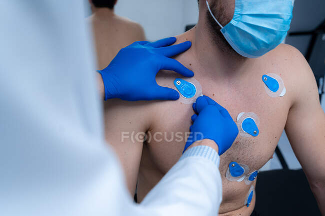 Médico de cultivo aplicando electrodos en el pecho de un paciente masculino irreconocible en máscara para la prueba de ECG - foto de stock