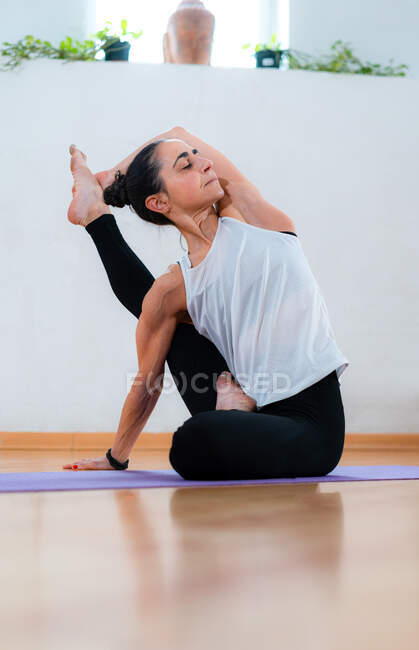 Рівень середнього віку жінка з закритими очима в спортивному одязі, що сидить на килимку для йоги і розтягує ногу в кімнаті — стокове фото