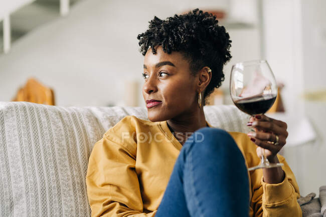 Content African American female sitting on sofa with glass of red wine and enjoying weekend at home while looking away — Stock Photo