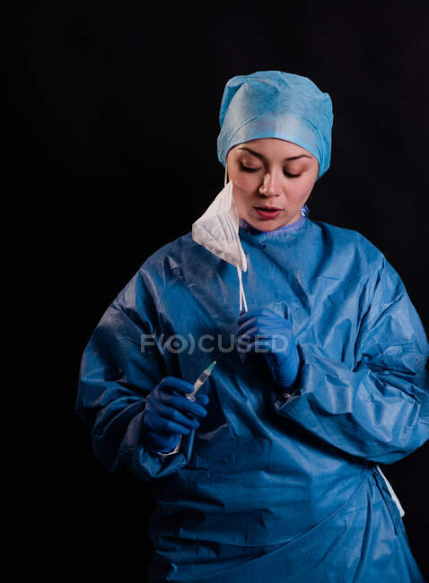 Mujer joven en uniforme médico quitándose las máscaras faciales mientras está de pie sobre fondo negro en la clínica - foto de stock