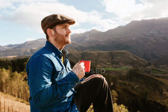 Side view of male hiker sitting on hill with metal cup of hot drink and admiring amazing scenery of mountainous terrain on sunny day — Stock Photo