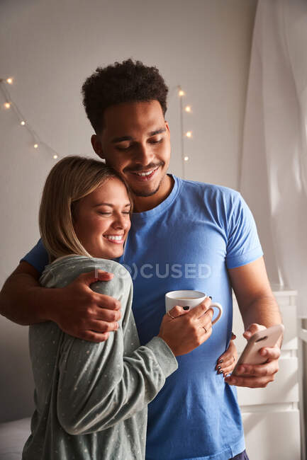 Side view of smiling multiracial couple in pajamas hugging in morning while taking selfie in morning at home — Stock Photo