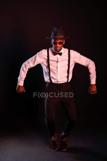 Jovem masculino étnico masculino modelo no chapéu e calças de pé dançando no titoes enquanto olhando para câmera no fundo preto — Fotografia de Stock
