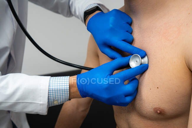 Médico masculino irreconocible recortado usando fonendoscopio mientras examina las embestidas de un paciente sin camisa en el hospital - foto de stock