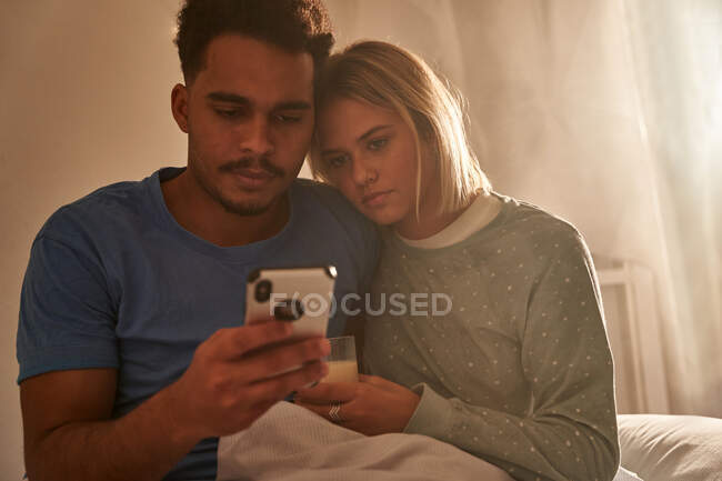 Calmo casal multirracial abraçando de manhã enquanto sentado na cama e usando o smartphone juntos — Fotografia de Stock
