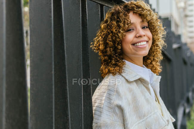 Vue latérale de charmante femme noire souriante aux cheveux bouclés appuyée sur une clôture métallique en ville et regardant la caméra — Photo de stock