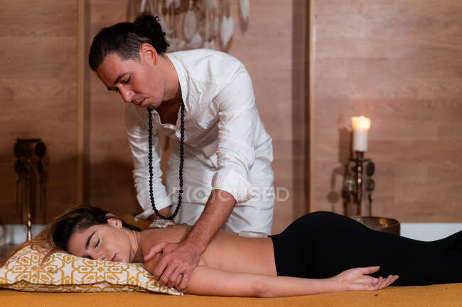 Мужчина-духовный терапевт массирует руку топлесс-женщины с закрытыми глазами, лежащей на подушке в помещении — стоковое фото