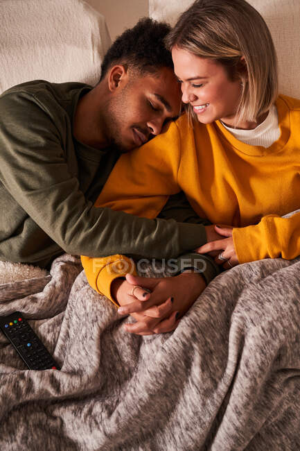 Angle élevé de couple multiethnique aimant se détendre sur le canapé sous la couverture tout en câlinant et en se tenant la main — Photo de stock