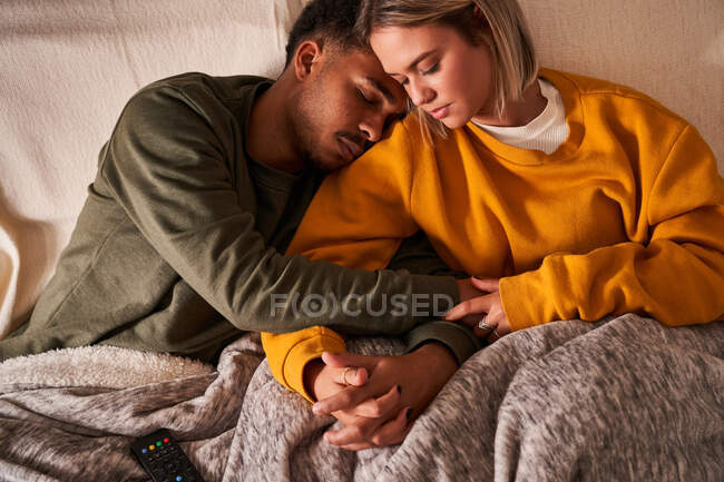 Alto angolo di amorevole coppia multietnica rilassarsi sul divano sotto coperta mentre coccole e tenersi per mano — Foto stock