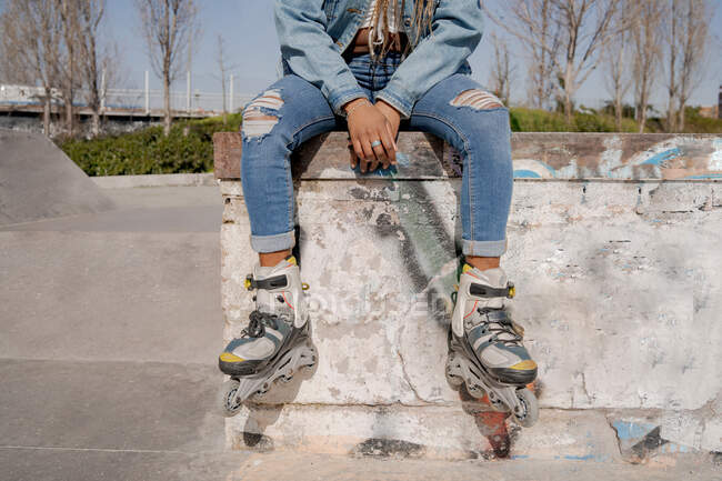 Crop femmina nera con acconciatura intrecciata e in pattini seduti su rampa in skate park e guardando altrove — Foto stock