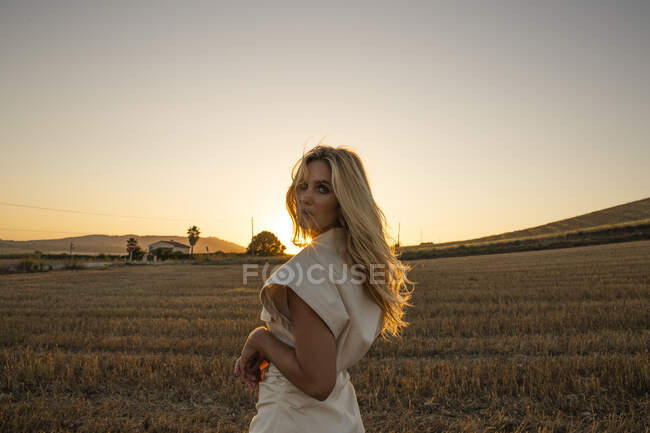 Peaceful female in elegant dress standing on dry field in rural area and looking at camera — Stock Photo