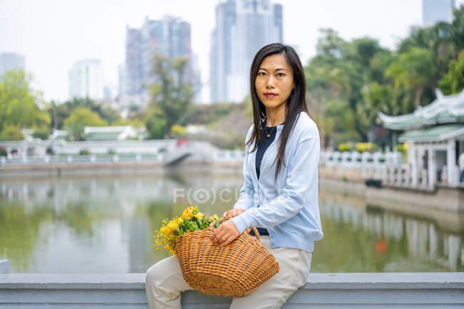Bella ragazza asiatica ritratto in un parco mentre si siede accanto a cesto di vimini con fiori gialli. — Foto stock