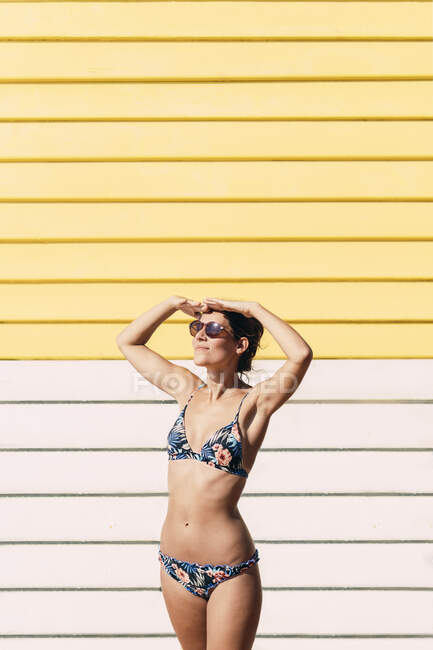 Cheerful female in trendy bikini and sunglasses standing on background of wooden bathing box and sunbathing in summer in Melbourne — Stock Photo