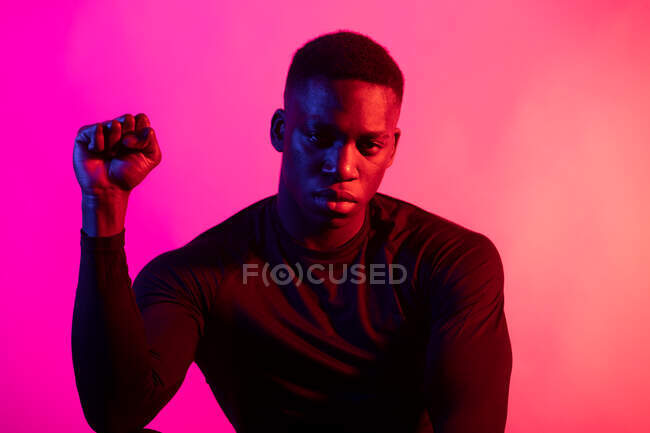 Fiducioso giovane nero in abito sportivo scuro guardando la fotocamera con le mani serrate a pugni su sfondo rosa fluo in studio scuro — Foto stock