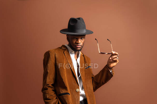 Joven hombre afroamericano en ropa de moda y sombrero mirando a la cámara sobre fondo marrón - foto de stock