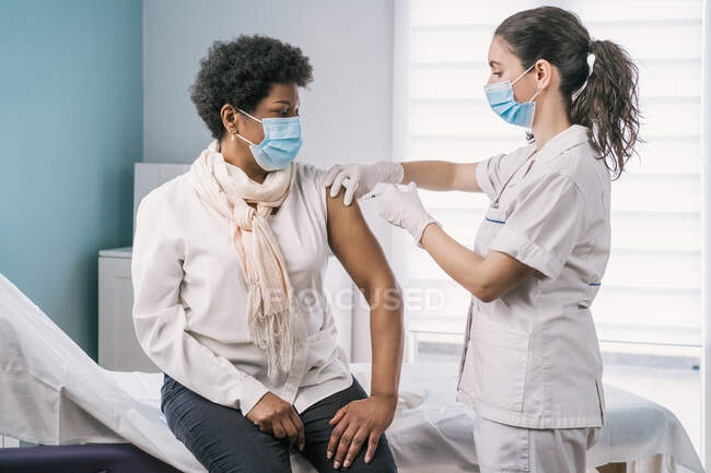 Жіночий медичний фахівець з захисної форми, латексні рукавички і особи з маскою вакцинують афроамериканського пацієнта в клініці під час спалаху коронавірусу. — стокове фото
