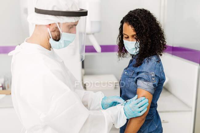 Seitenansicht eines männlichen Facharztes in Schutzuniform, Latexhandschuhen und Gesichtsschutzimpfung einer afroamerikanischen Patientin in der Klinik während des Coronavirus-Ausbruchs — Stockfoto