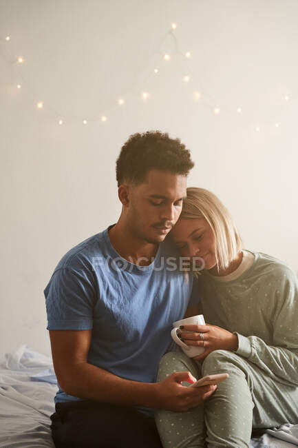 Lächelndes multiethnisches Paar im Schlafanzug, das sich morgens umarmt, während es morgens zu Hause auf dem Smartphone surft — Stockfoto