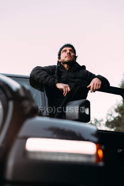 De baixo aventureiro inclinado na porta de um carro off-road, enquanto olha para longe antes de iniciar a viagem — Fotografia de Stock